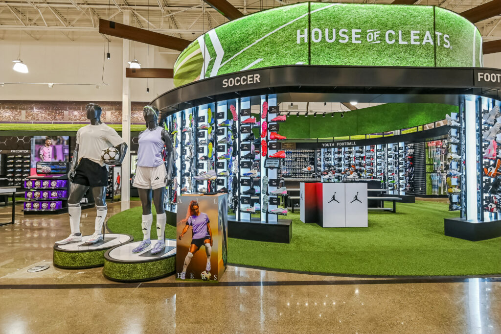 A “House of Cleats” soccer retail display featuring two mannequins in athletic apparel, rows of soccer cleats on wall shelves, and an artificial grass floor.