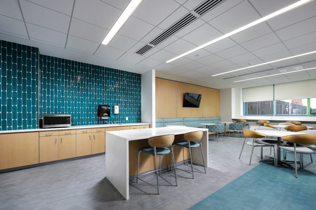 A modern office breakroom with a teal patterned feature wall, wood cabinets, a white kitchen island with two bar stools, and several dining tables with chairs.