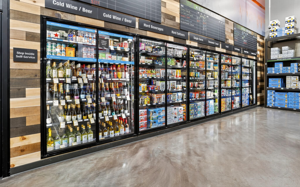 A wide refrigerated display case in a store, stocked with various alcoholic and non-alcoholic beverages, with overhead signage labeled by category and a keg menu board.