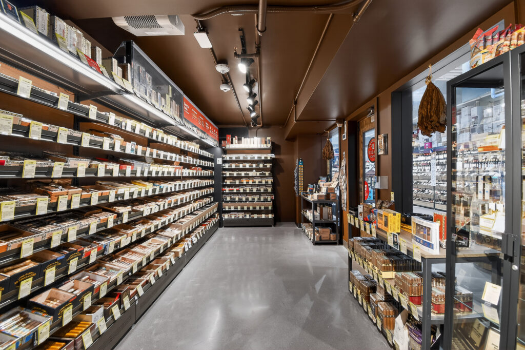 Interior of a cigar shop with long shelves displaying rows of boxed cigars, a center display unit, and a glass-enclosed humidor.