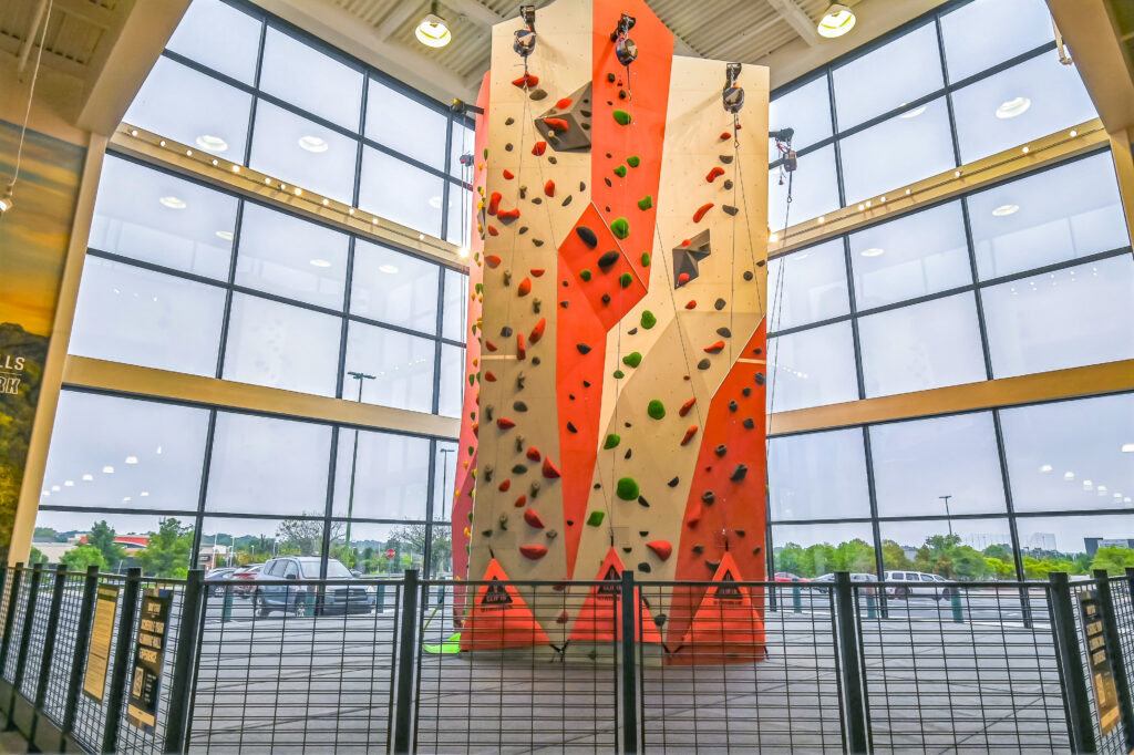 Indoor rock climbing tower with orange and tan panels, situated in front of a large windowed wall.
