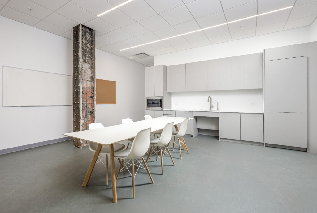 A modern office kitchenette with light grey cabinetry, a stainless steel sink, and a long white table with six white chairs.