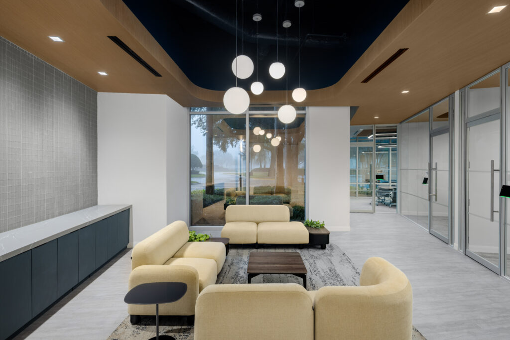 Modern office lobby with three beige sofas arranged around a coffee table, illuminated by spherical pendant lights under a dark ceiling cutout.