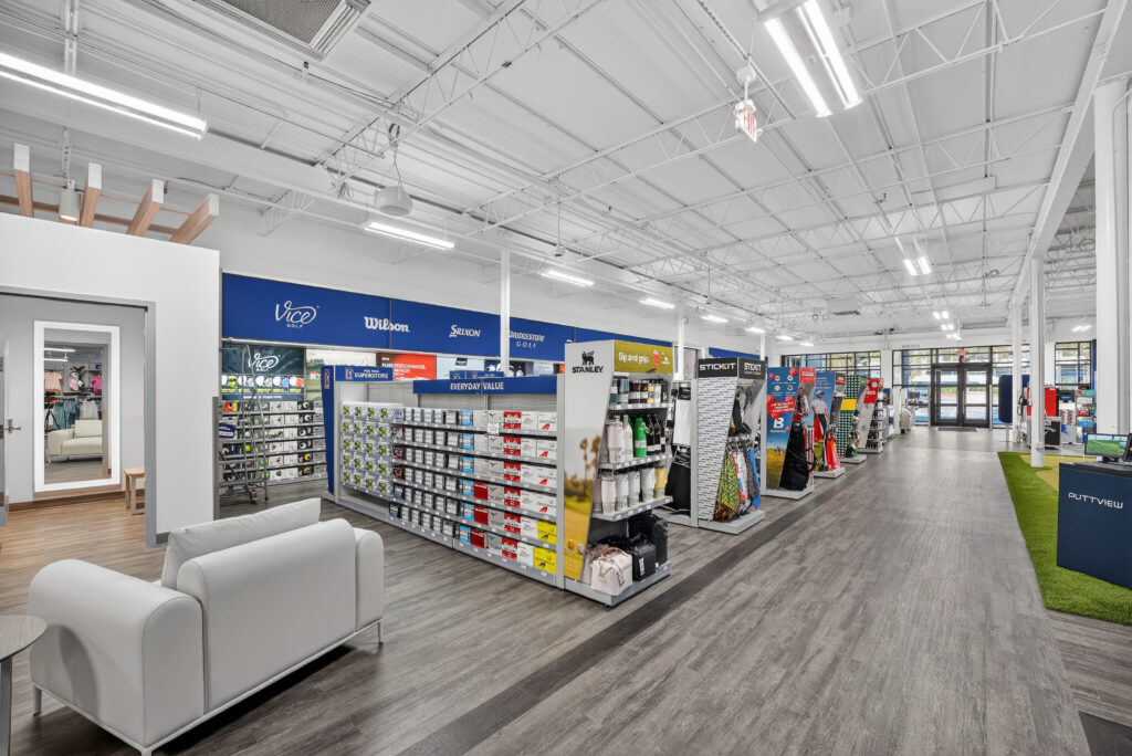 Interior of a golf retail store with rows of shelves displaying golf balls, equipment, and accessories, and a white sofa in the foreground.