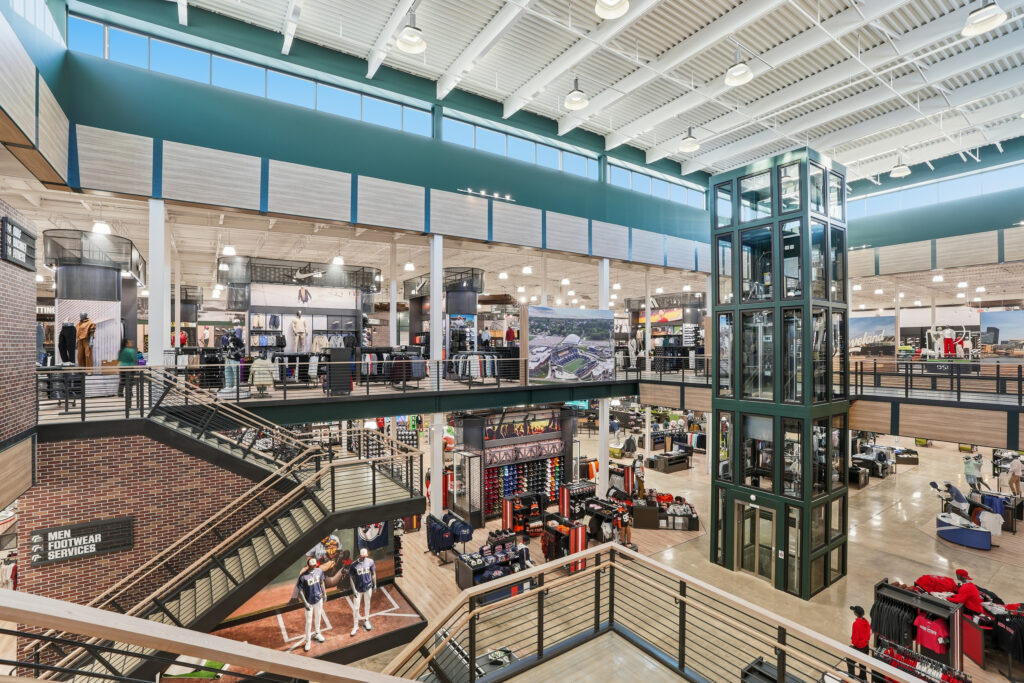 Interior of a multi-level sporting goods store with a central glass elevator, stairs, and various apparel displays.