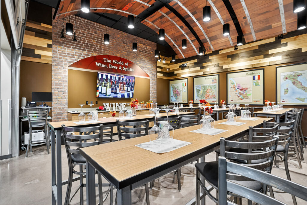 Interior of a wine tasting room featuring long wooden tables with metal chairs, wine glasses, and maps of wine regions on the brick walls.