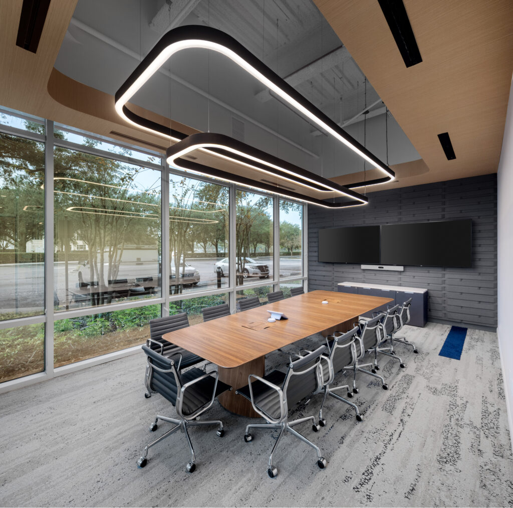 A modern conference room with a long wood table, swivel office chairs, and two horizontal pendant lights under a wooden ceiling panel.