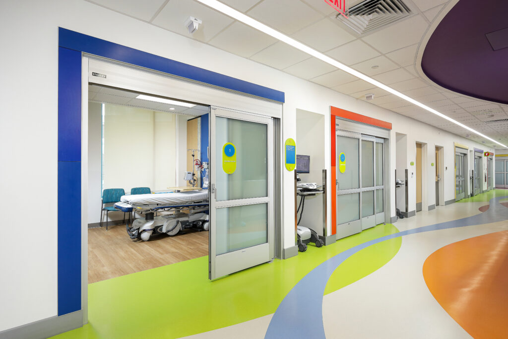A bright hospital hallway with sliding doors opening into an exam room containing a medical bed and blue chairs.