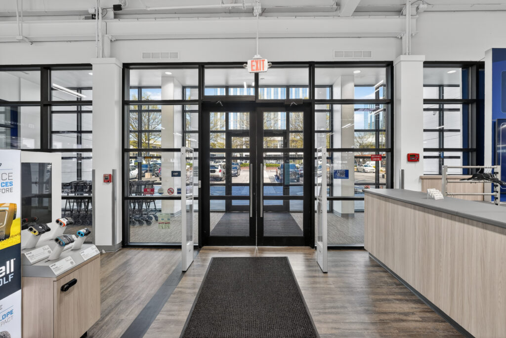 Glass double doors at the entrance of a retail store with a dark floor mat, an illuminated exit sign, and a customer service desk to the right.