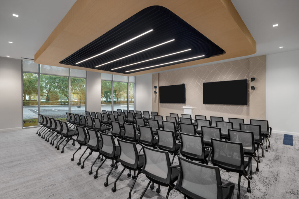 Rows of black mesh office chairs face a lectern and two wall-mounted televisions in a conference room with floor-to-ceiling windows.