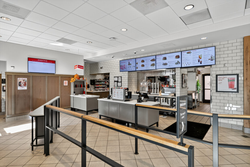 Interior of a fast-food restaurant showing the service counter, digital menu boards, and a waiting area with metal railings.