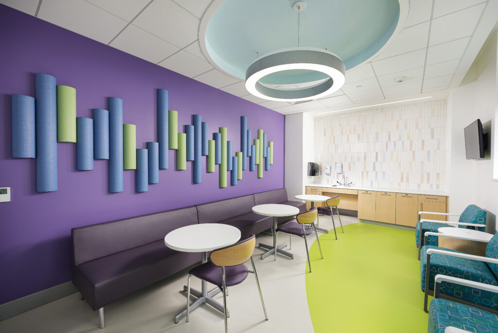 A modern lounge area with a purple wall featuring cylindrical acoustic panels, a long banquette, round tables, and seating with teal patterned cushions.
