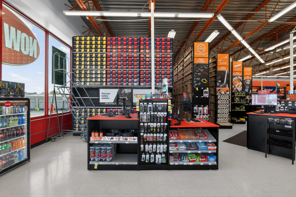 Interior of an auto parts store showing shelves of batteries, various merchandise, a checkout counter with snacks, and branded signage.