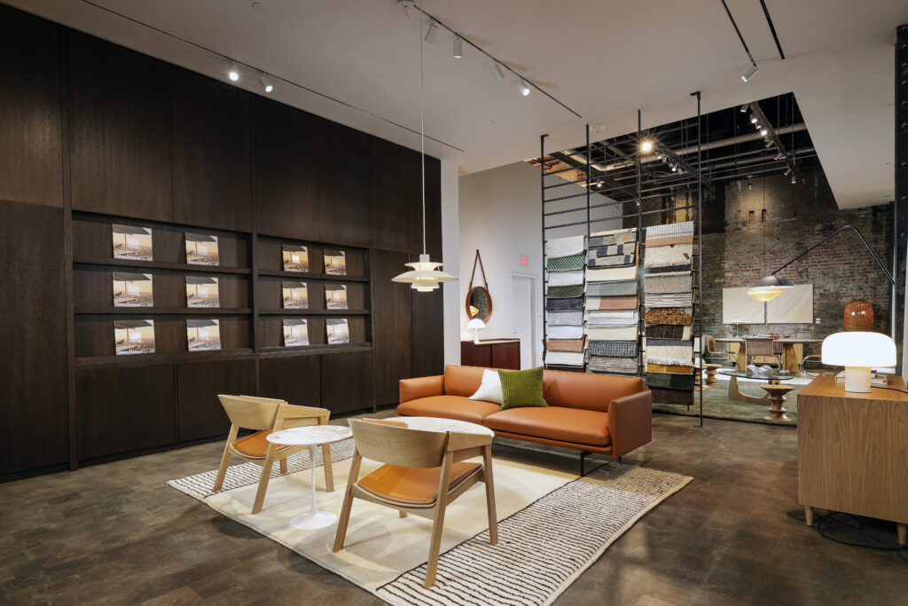 Furniture showroom featuring a leather sofa and two wooden chairs on a patterned rug, with shelving displays, fabric samples, and home accessories in the background.