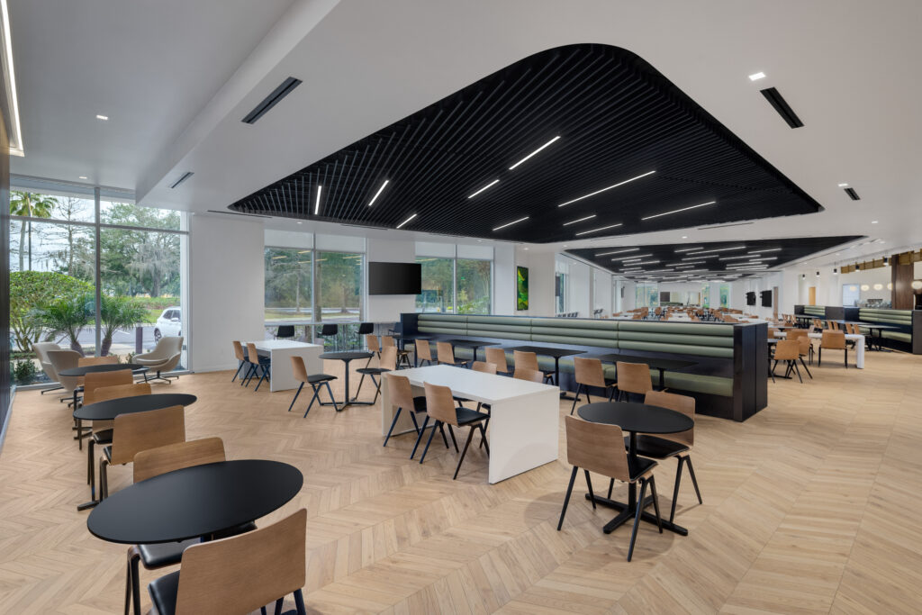 Modern office cafeteria with rows of round and rectangular tables, wooden chairs, and a long green banquette seating area.