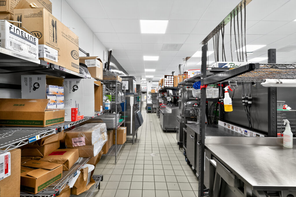 A commercial kitchen aisle with metal shelving units stacked with cardboard boxes and a stainless steel prep station.