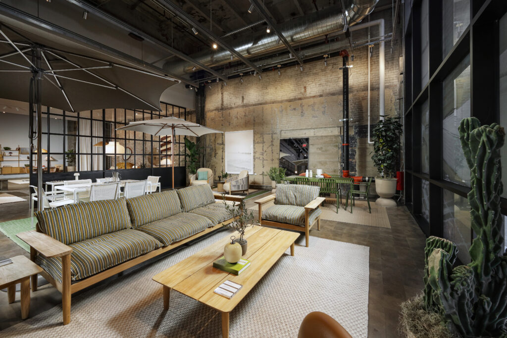 A modern furniture showroom featuring a striped sofa and matching armchair, a wooden coffee table, and an adjacent dining area with a green table and chairs in an industrial space with exposed brick walls.