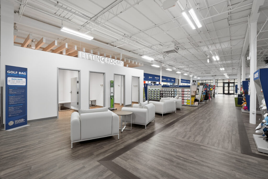 Golf store interior with a row of fitting rooms, white waiting area chairs, and aisles displaying golf bags and equipment.
