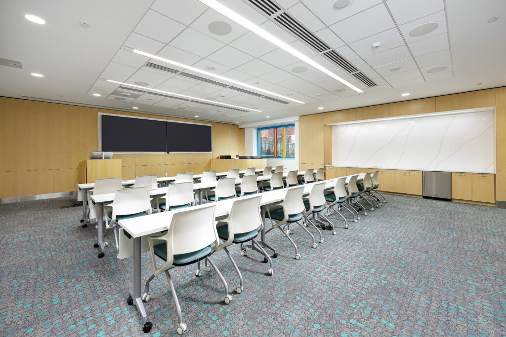 A classroom with rows of white chairs and tables, featuring a large black display screen at the front and a white marker board along the side wall.