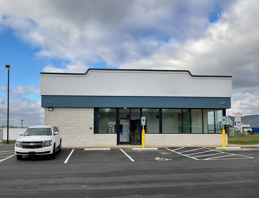 A vacant commercial building with a white and blue exterior and a large front window, with a white SUV parked in the adjacent lot.