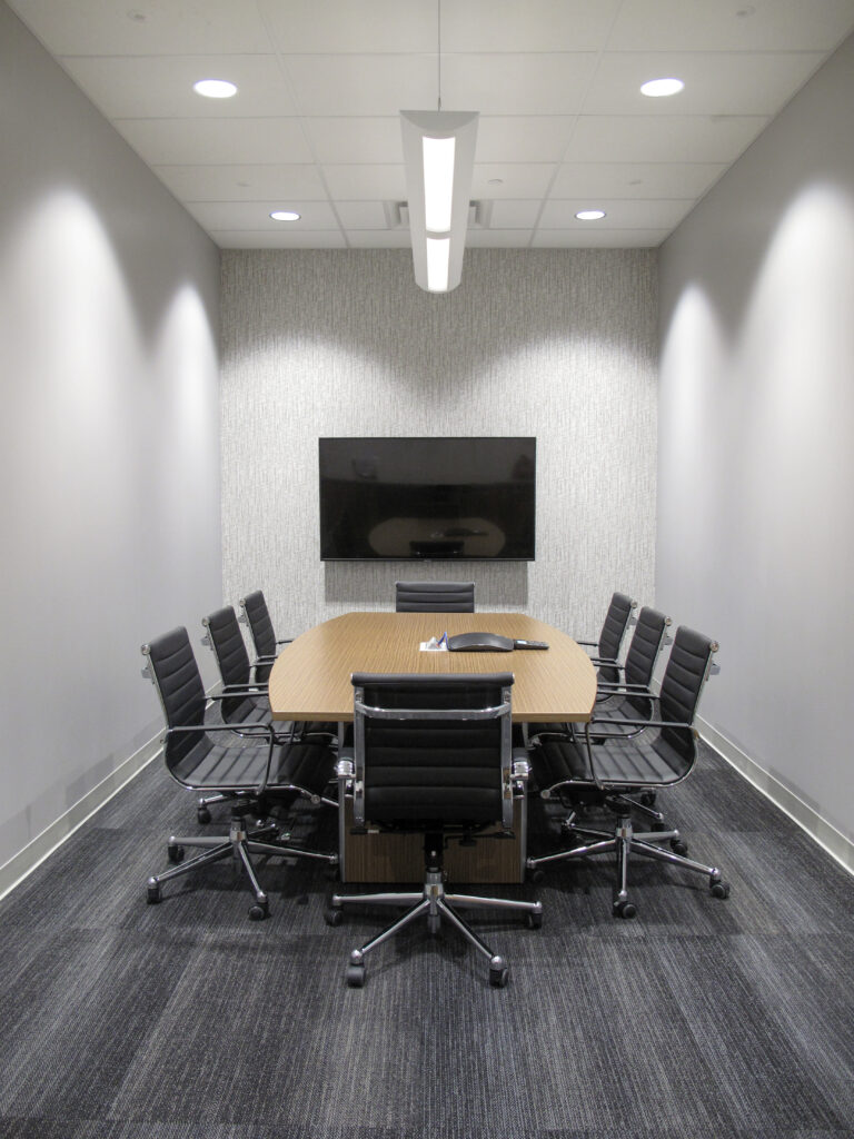 A small conference room with an oval wooden table, eight black rolling chairs, and a wall-mounted television.
