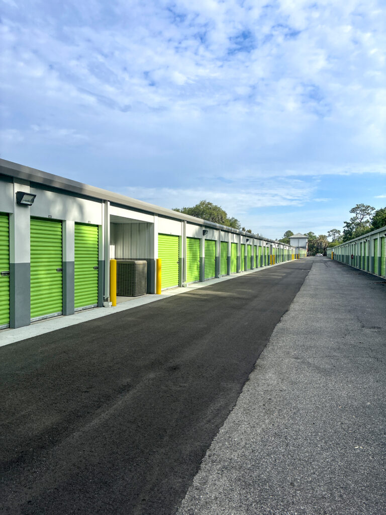 A row of storage units with green roll-up doors along a paved drive.