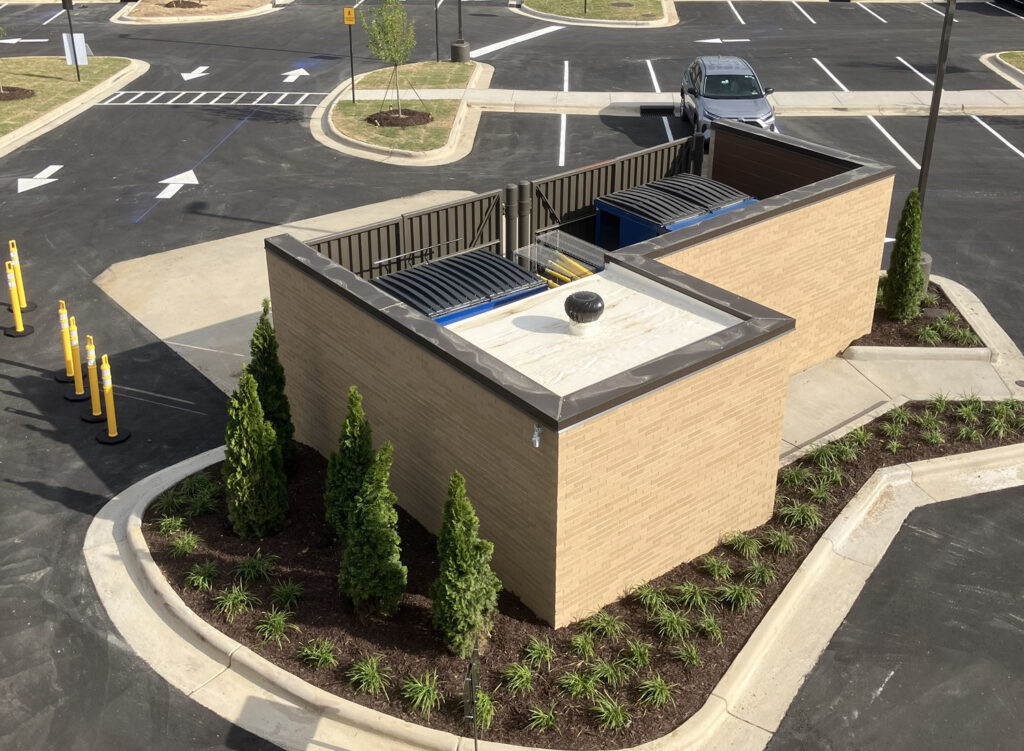 High-angle view of a brick dumpster enclosure in a parking lot, with two dumpsters inside and landscaping along the front.