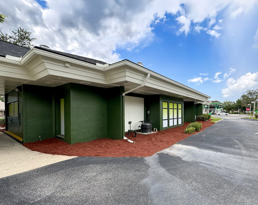 The exterior of a dark green commercial building with a white roofline, surrounded by red mulch and an asphalt parking lot.