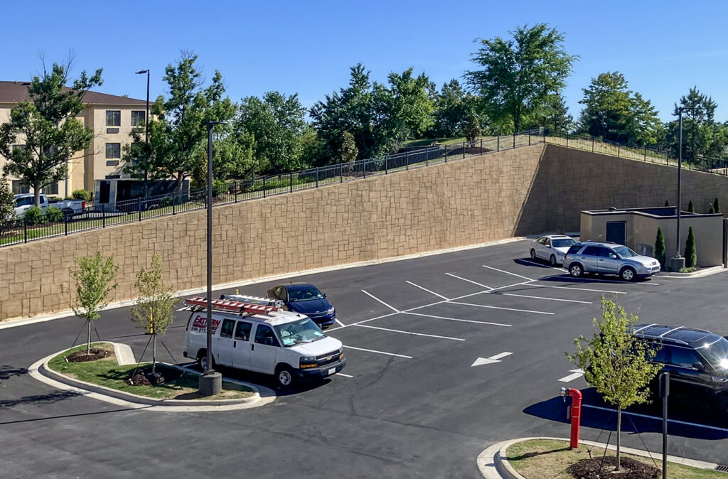 A parking lot with a light-colored commercial van, a dark blue sedan, a silver SUV, and a black SUV, situated below a tiered landscape wall.