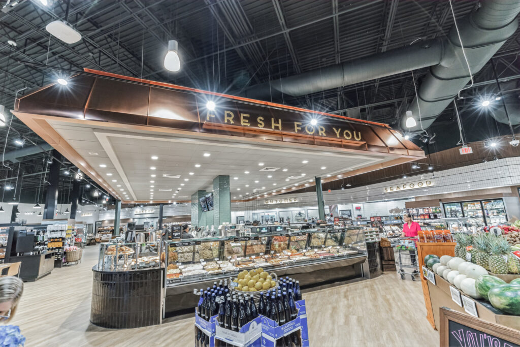 Grocery store interior featuring a central “Fresh For You” service counter with displays of prepared foods, bakery, and meat, with produce in the foreground.