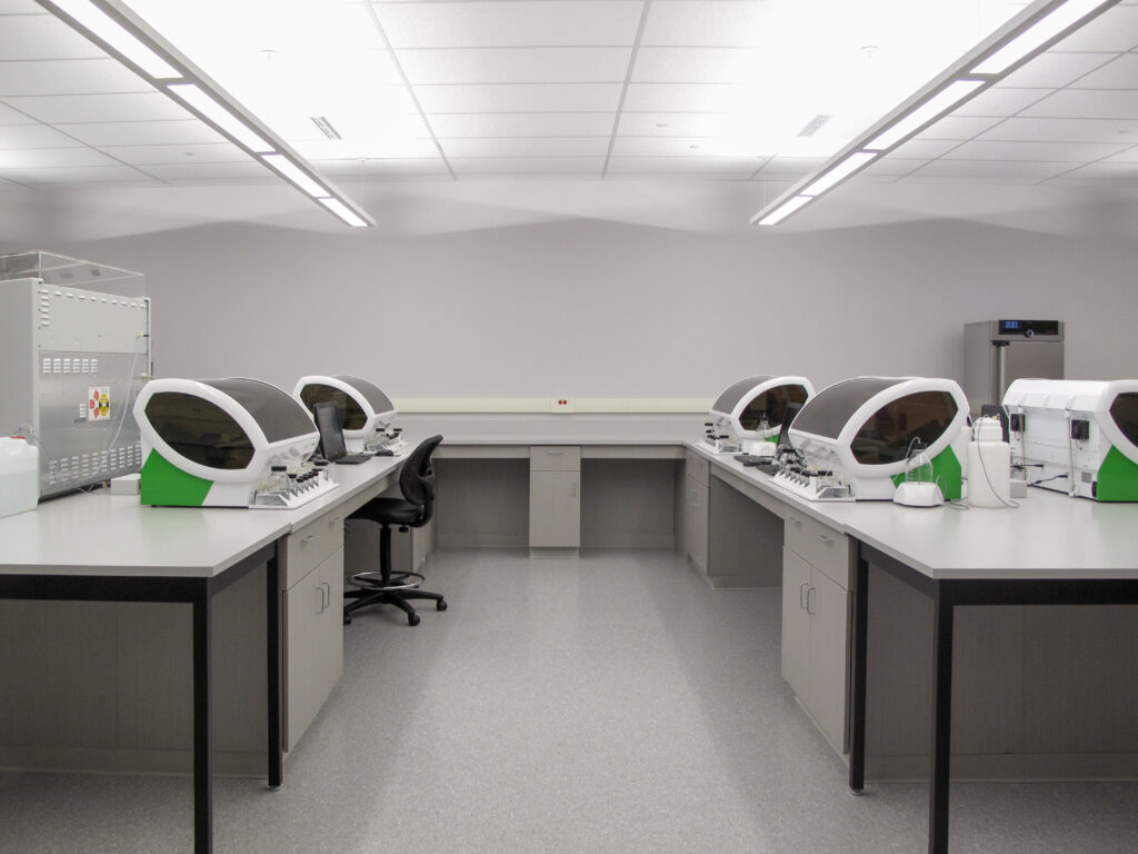 A laboratory workspace with rows of automated diagnostic analyzers on grey workbenches.
