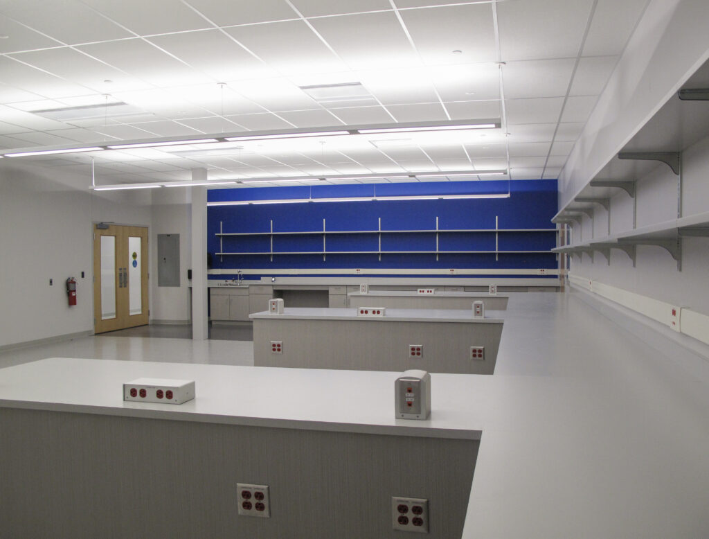 An empty science laboratory with white workbenches, electrical outlets, and blue wall-mounted shelving.