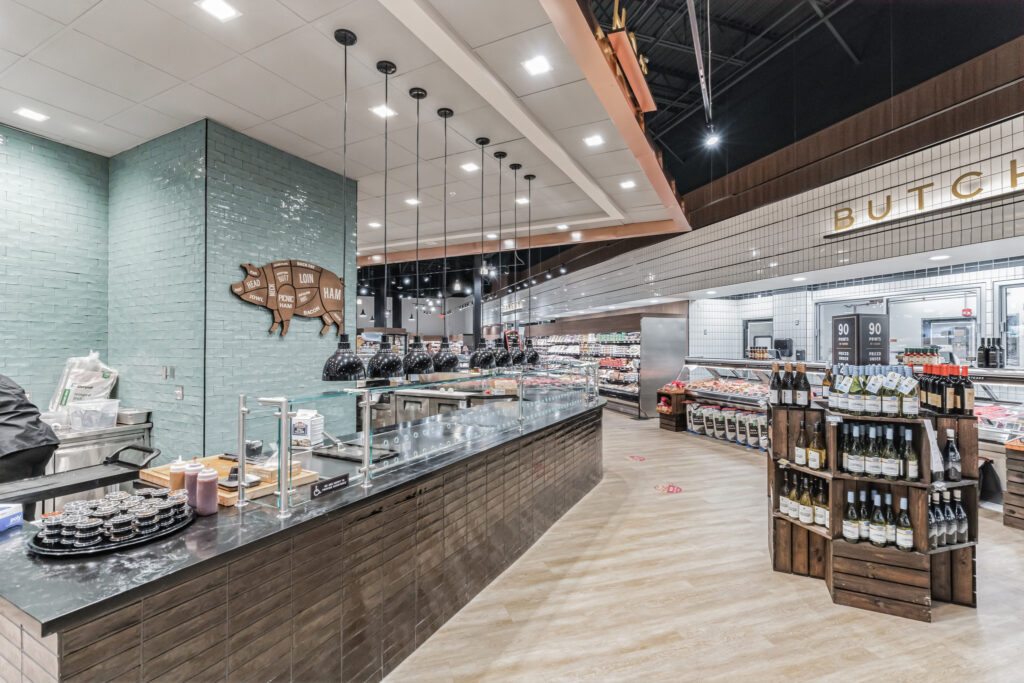 A grocery store butcher counter with a pork diagram on a teal tiled wall, black pendant lights, and a wooden rack of wine bottles in the foreground.