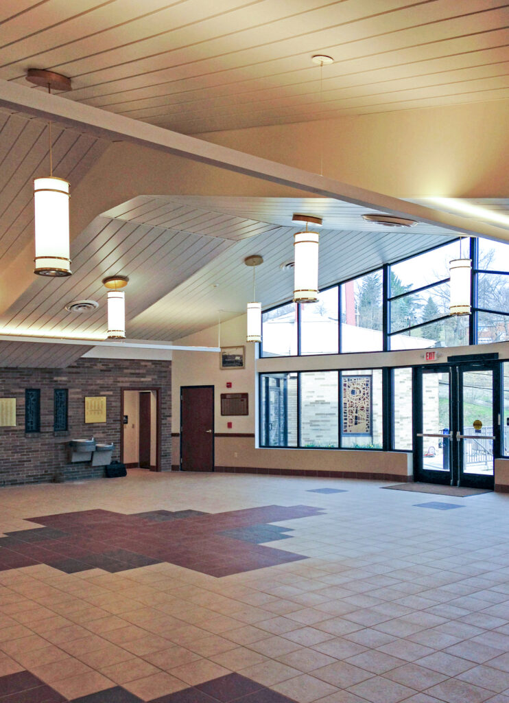 A spacious, empty lobby with tile floors, hanging pendant lights, and large windows looking out to a tree-lined street.