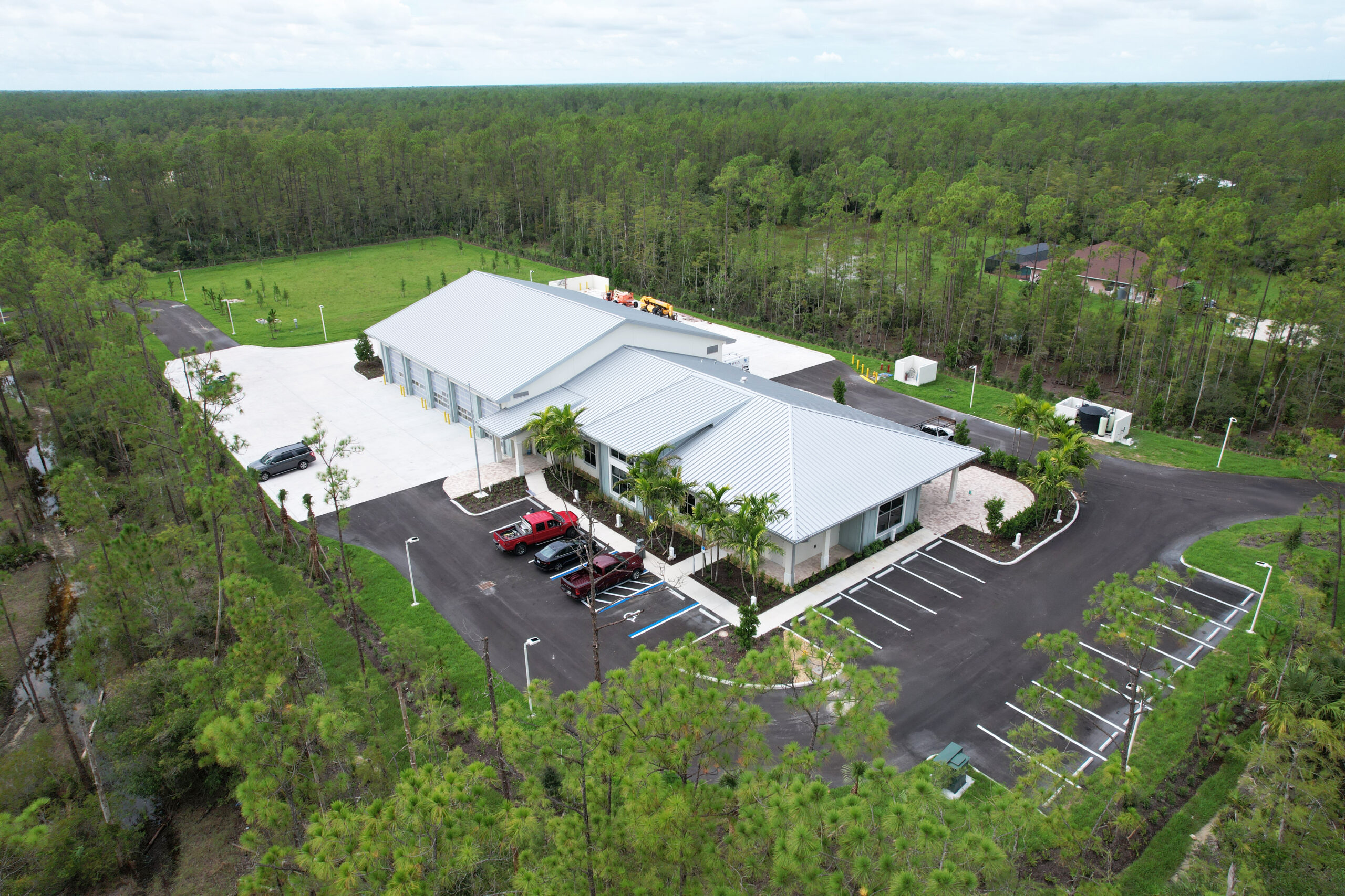 Aerial view of a commercial building with a metal roof, parking lot, and surrounding pine forest.