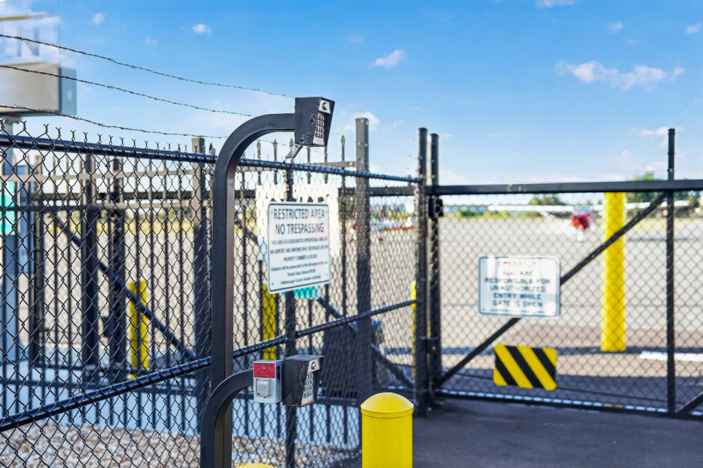 A chain-link gate at an airfield entrance with a keypad, a red emergency button, and “Restricted Area” signage.
