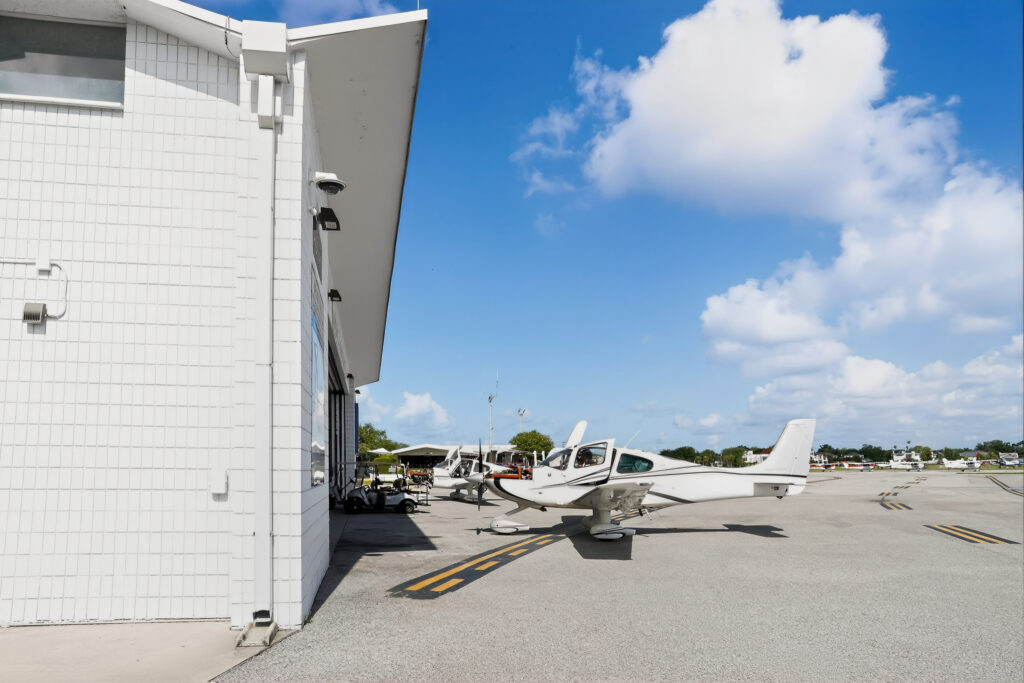 Small white airplane parked on an airport tarmac next to a hangar building.