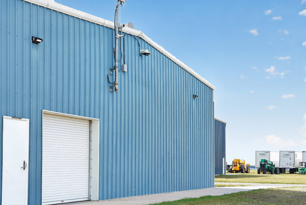 Blue corrugated metal industrial building with a roll-up garage door and security equipment mounted on the exterior wall.