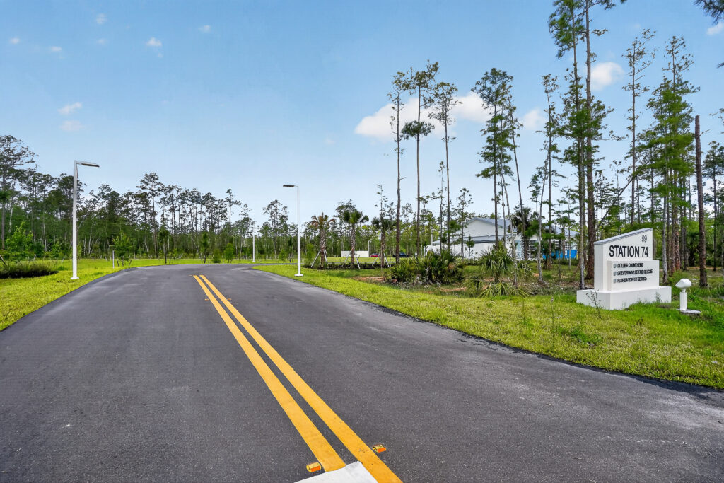 An asphalt road leads to a sign for “Station 74” serving Collier County EMS, Greater Naples Fire Rescue, and Florida Forest Service, set in a wooded area.