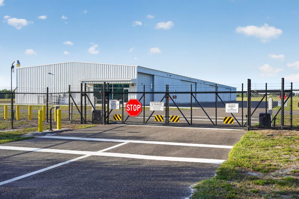 A large metal warehouse building behind a black chain-link fence with a stop sign and restricted area signs on the gate.