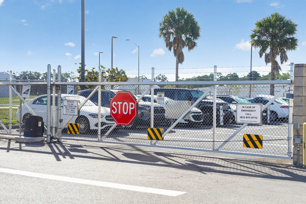 A closed chain-link gate with a stop sign and two informational signs in front of a parking lot containing multiple cars and two palm trees.
