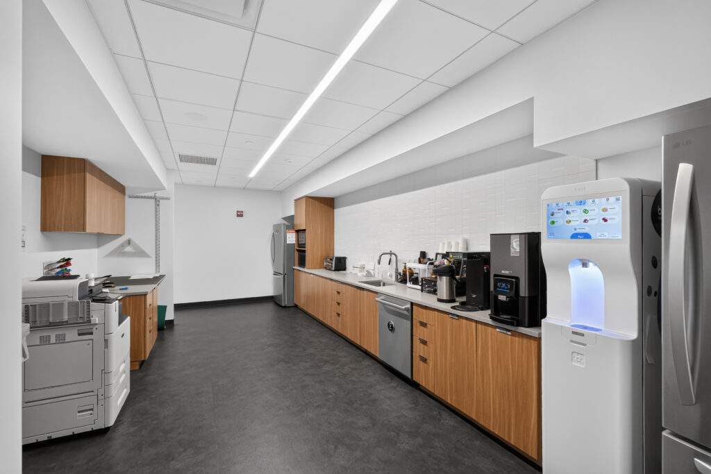 An office breakroom featuring a long counter with a sink, coffee machines, a dishwasher, and a water dispenser, with a printer on the left.