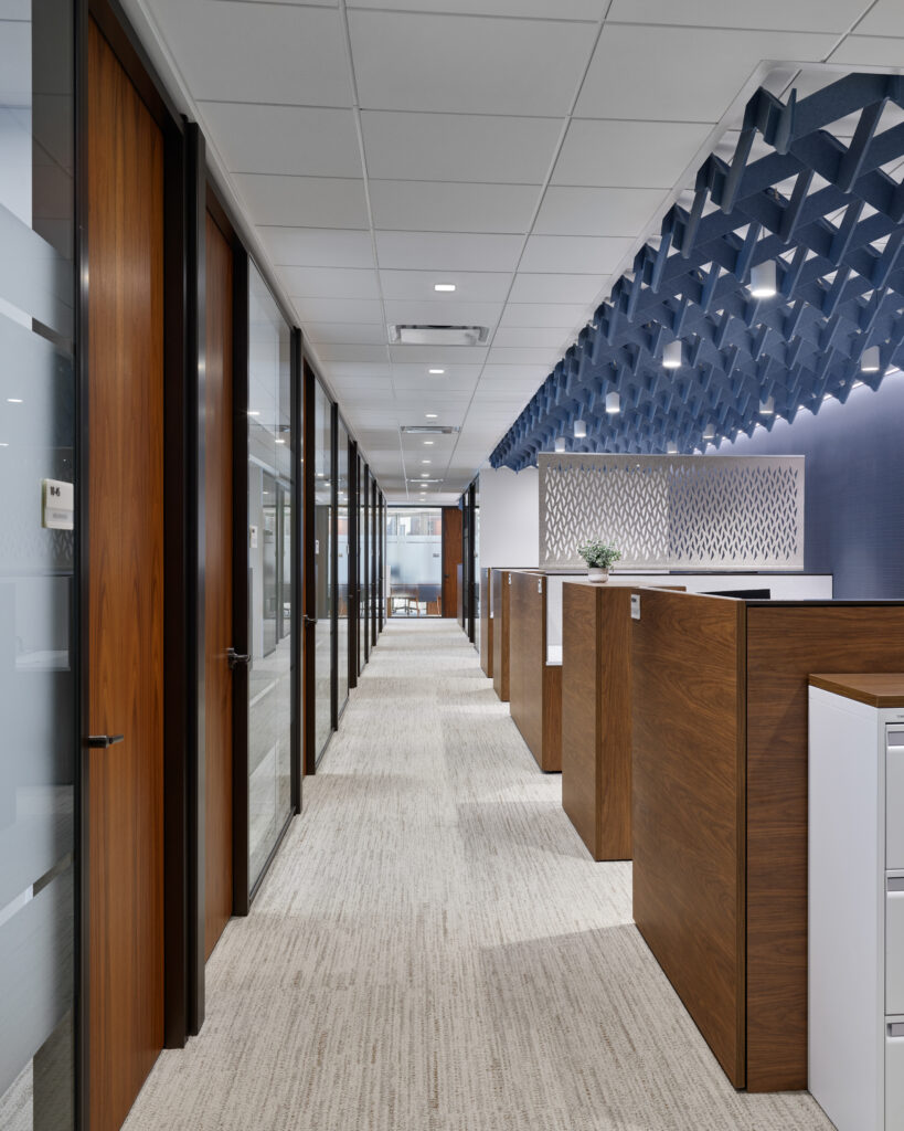 Office hallway with glass-fronted offices on the left and wooden cubicle partitions on the right, under a geometric blue ceiling feature.