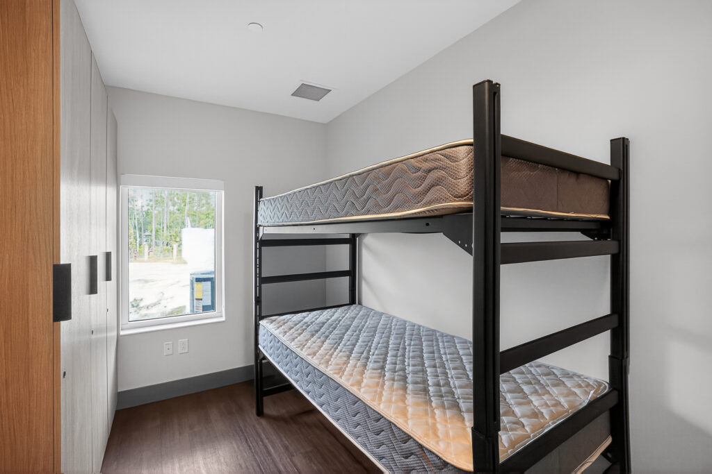 A black metal bunk bed with two mattresses in a minimalist room with a window looking out onto a wooded area.