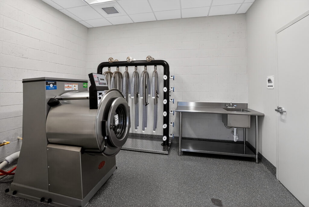 Industrial washing machine next to a wall-mounted rack for hanging items and a stainless steel utility sink.