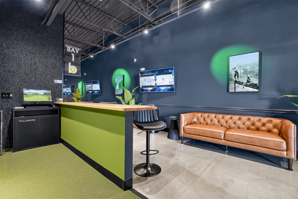 Indoor golf simulator bay featuring a monitor, a high-top counter with a stool, and a brown leather sofa.