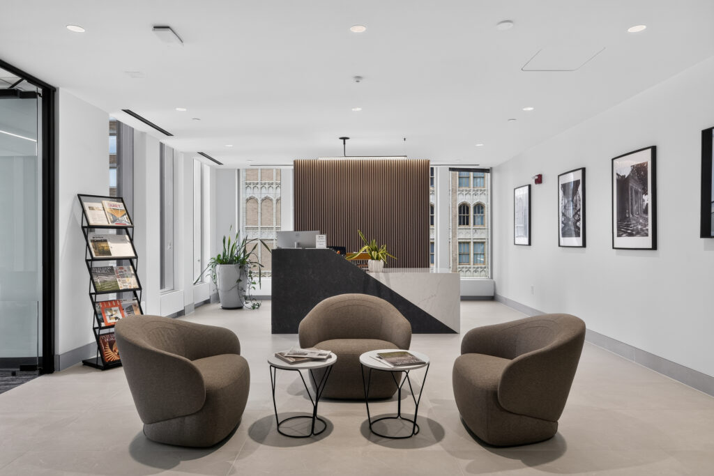 A modern office lobby with three brown lounge chairs, two small circular tables, and a reception desk featuring a dark stone base and wood-paneled backdrop.