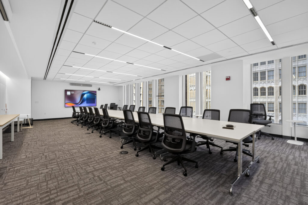 A long conference table surrounded by black ergonomic chairs in a brightly lit office with large windows.