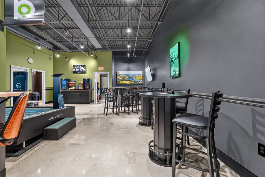 Interior of a golf lounge featuring putting greens, high-top tables with chairs, a refreshments area, and wall-mounted televisions.