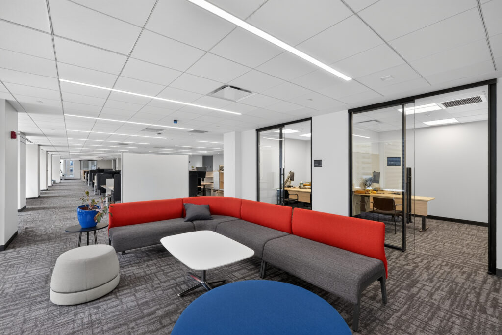 A lounge area in an office with a red and gray modular sofa, a white coffee table, and a blue ottoman, adjacent to two glass-walled private offices.
